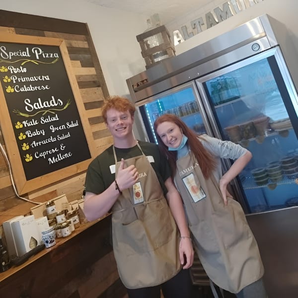 Two smiling Altamura Pizza employees in aprons stand by a menu board and a refrigerator.