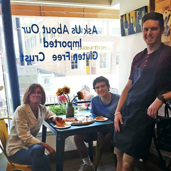 Three people, two seated and one standing, enjoying Altamura Pizza with slices on plates.