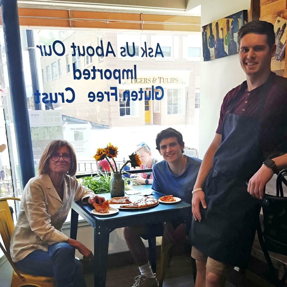 Three people, two seated and one standing, enjoying Altamura Pizza with slices on plates.