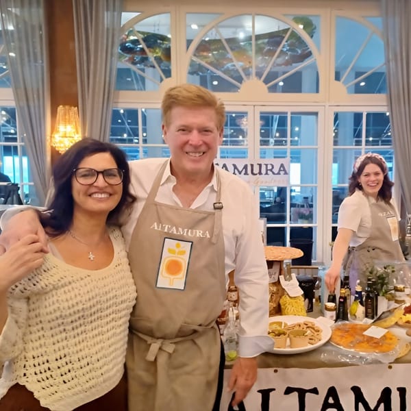 Two smiling people, one wearing an Altamura apron, stand by a table of Altamura Pizza products.