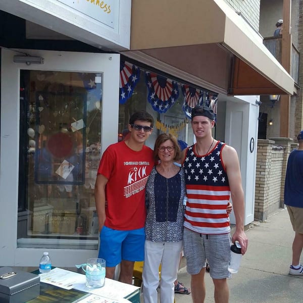 Three people, two young men and a woman, stand smiling outside a storefront decorated with patriotic bunting.