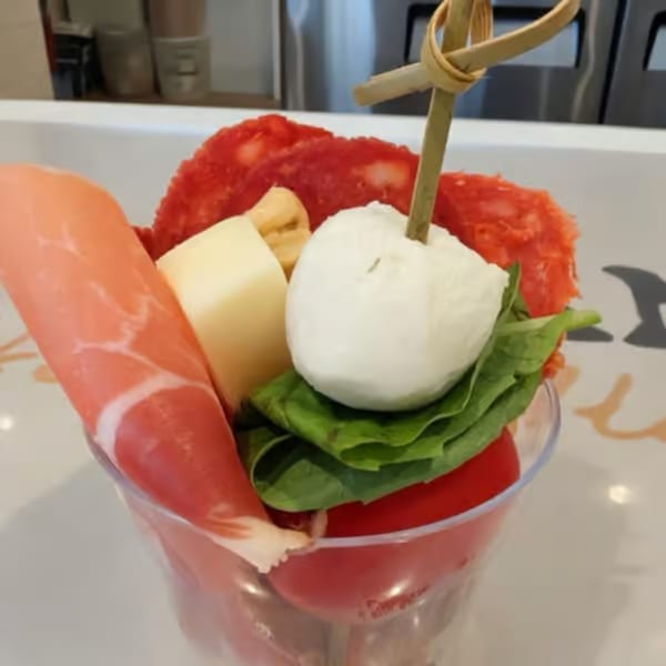 A clear cup filled with prosciutto, salami, cheese, mozzarella, basil, and tomatoes.
