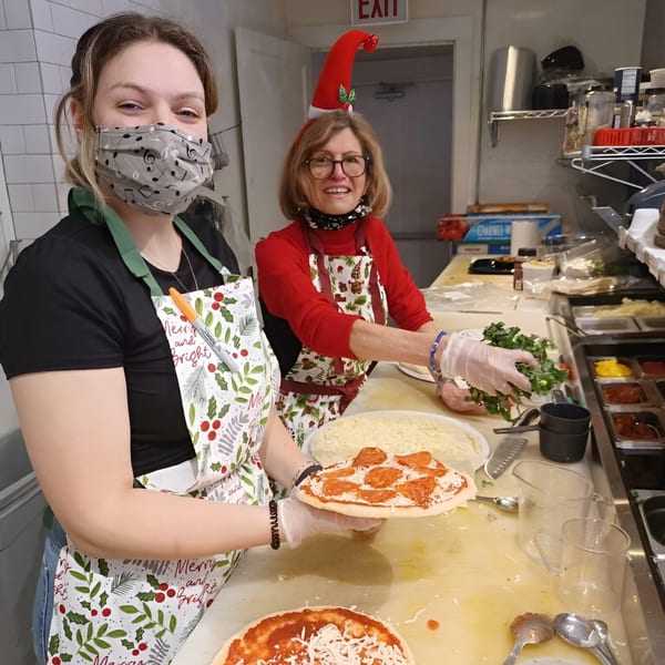 Two women in festive aprons prepare pizzas in a kitchen, one holding a pepperoni pizza.