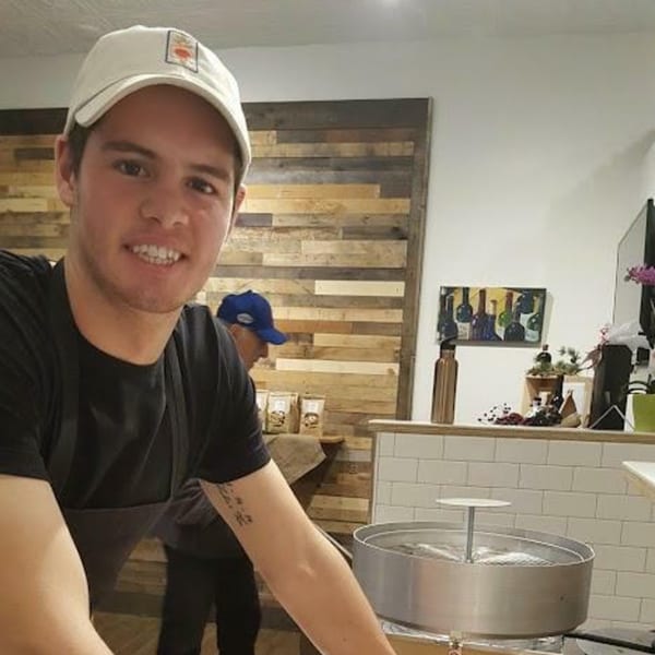 Smiling young man in a white cap and black apron stands in a rustic-chic kitchen.