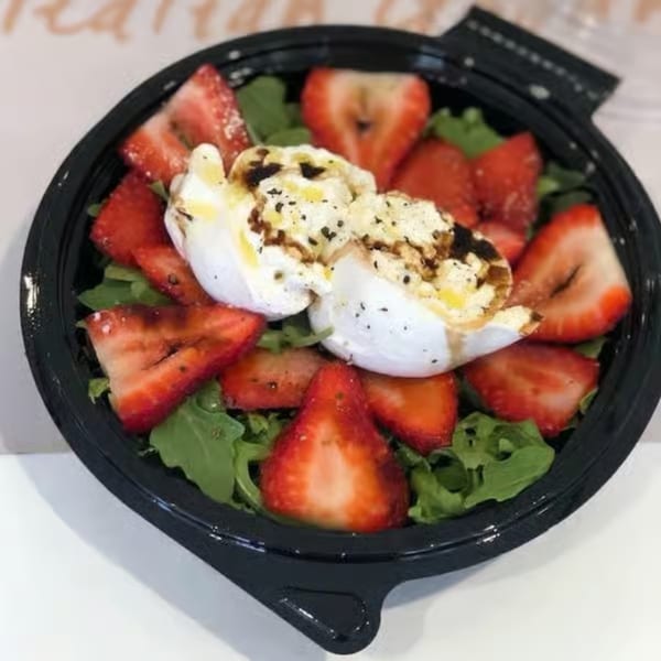 A black bowl filled with fresh arugula, sliced strawberries, and creamy burrata cheese drizzled with balsamic glaze.