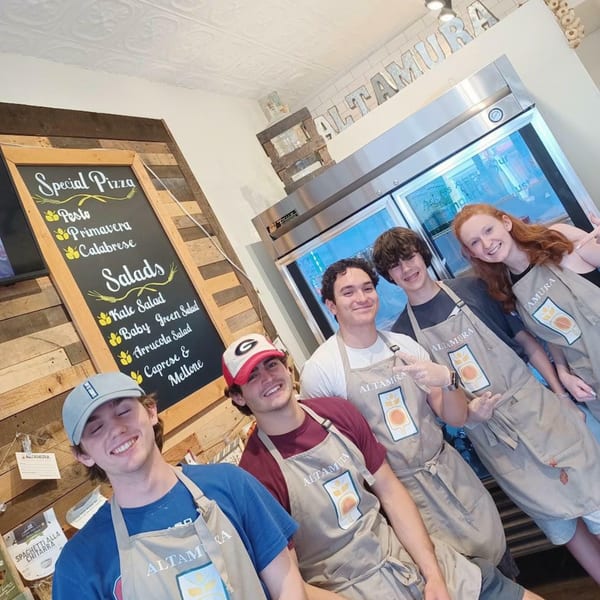 Five smiling young Altamura staff members in aprons stand in front of a menu board and refrigerator.