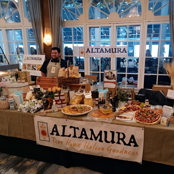 Altamura Pizza vendor smiling behind a table laden with Italian food products and pizzas.
