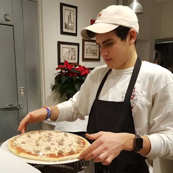 A young man in an apron and cap prepares an Altamura Pizza with cheese and sausage.