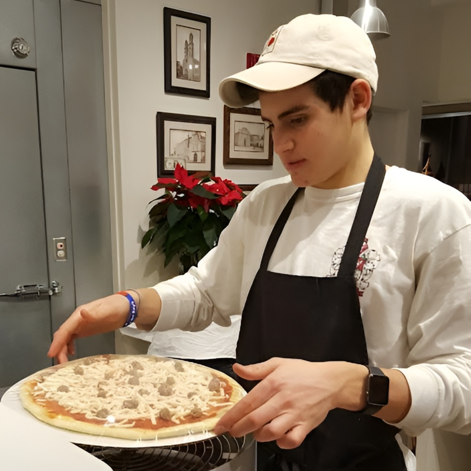 A young man in an apron and cap prepares an Altamura Pizza with cheese and sausage.