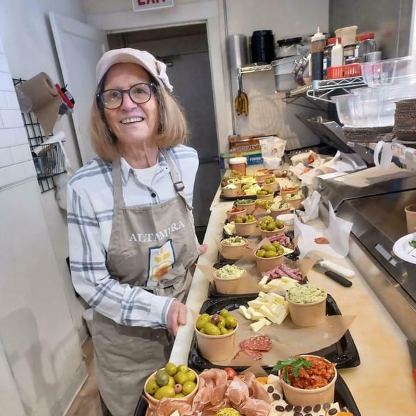 A smiling woman in an Altamura apron stands behind a counter filled with charcuterie boards.